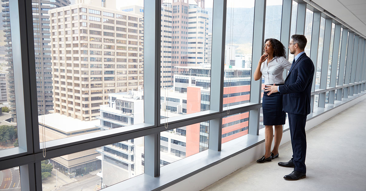 Woman and man in business attire standing in a large empty office, looking out a large window at a downtown city skyline.