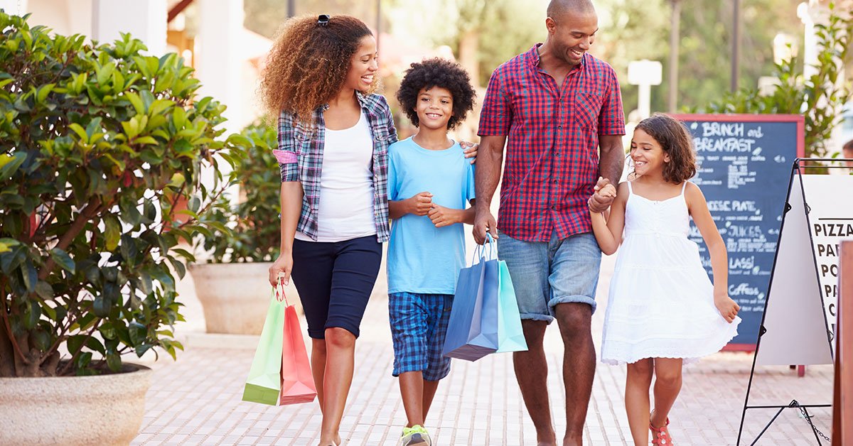 A family, including a mother, father, son and daughter, are walking outside in a shopping plaza. The parents hold shopping bags.