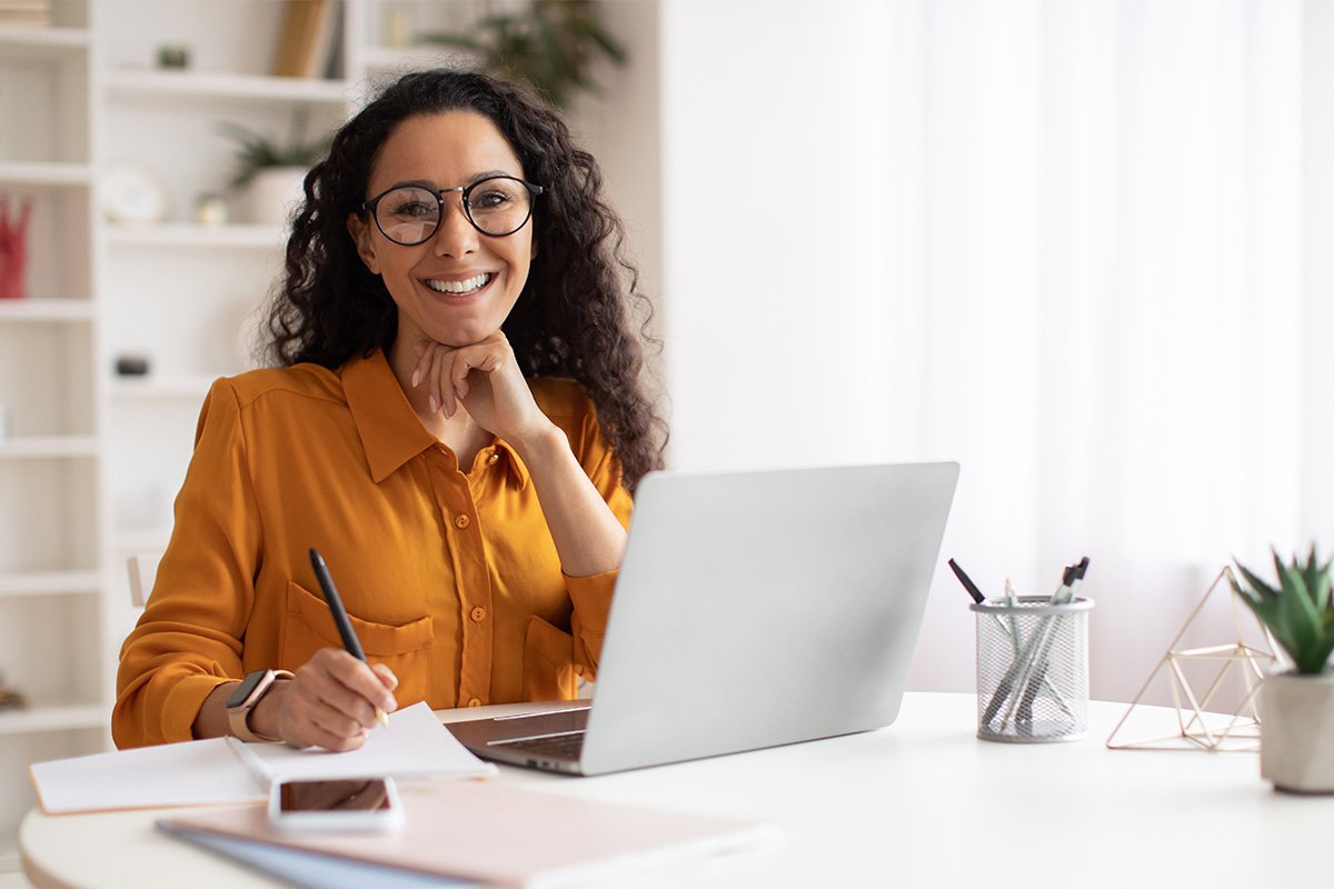 Woman with smiling and sitting at a desk with a laptop and a pen in her hand 