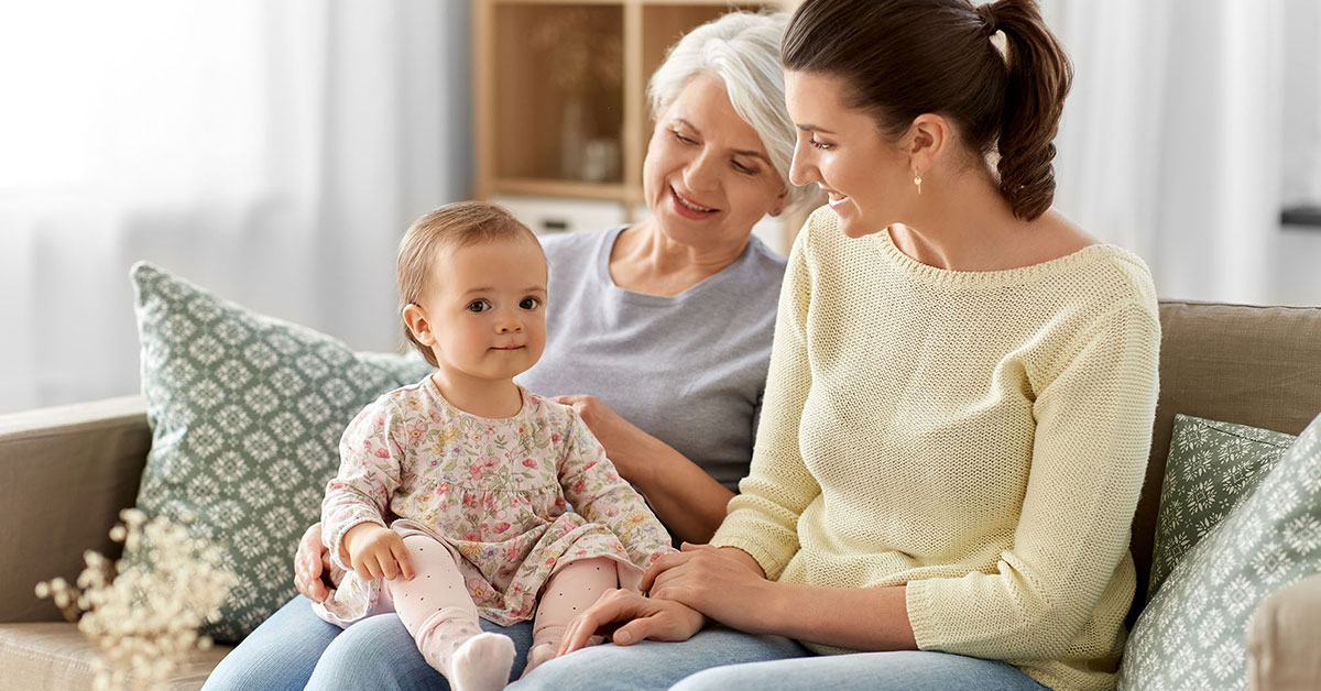 Granddaughter sits on her grandmother's lap as her mom sits next to them and smiles down at her.