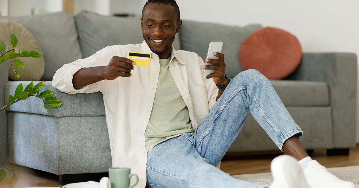 Man seated in a living room against a couch, activating his new checking account debit card while holding a mobile phone.