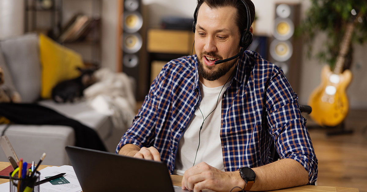 Self-employed man seated at table working on laptop while talking with client through headset