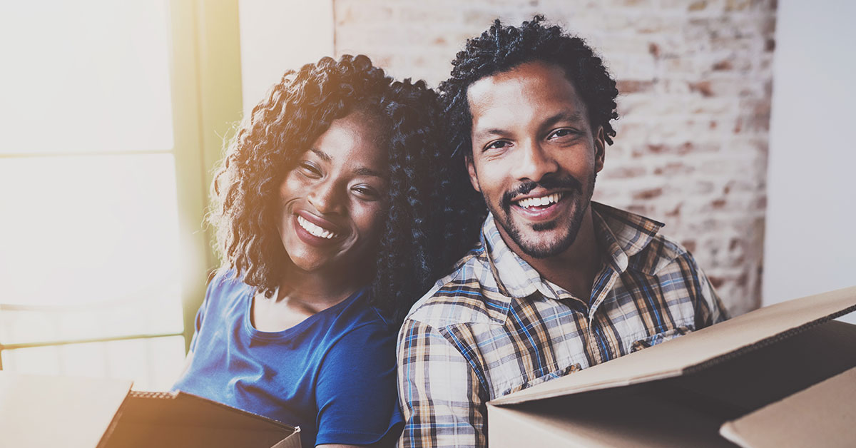 Close up photo of young couple smiling and looking at the camera. They are holding cardboard boxes, indicating they are moving into a new home.