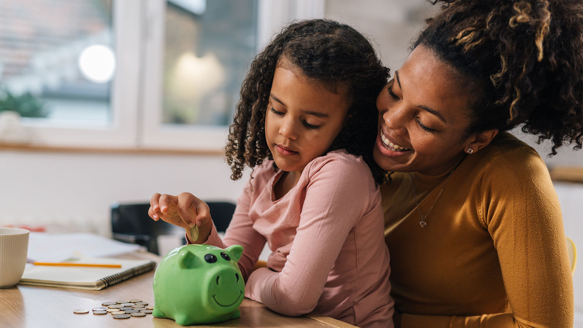 A mom smiles as she teaches her daughter how to save money by putting coins in a piggy bank.
