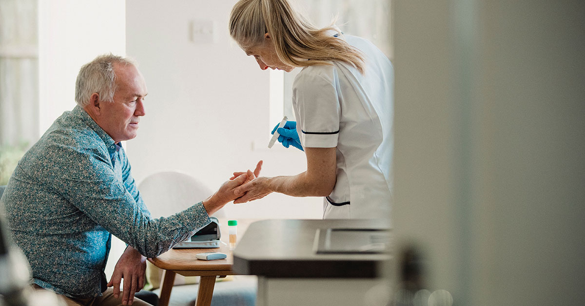 An older man sits at a table in his kitchen. A nurse is holding his finger, taking a blood sample to check his blood sugar