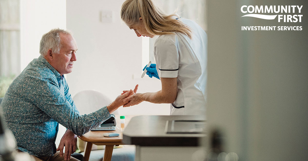 An older man sits at a table in his kitchen. A nurse is holding his finger, taking a blood sample to check his blood sugar