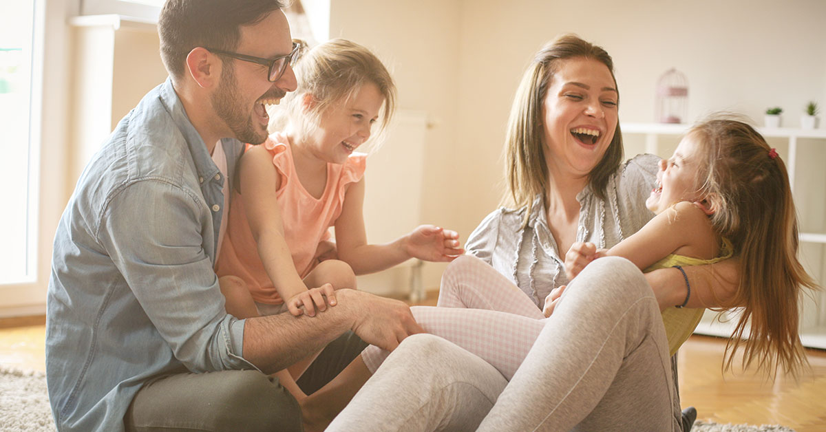 A happy family consisting of a mom, dad, and two daughters, sitting on the floor in their home, laughing together