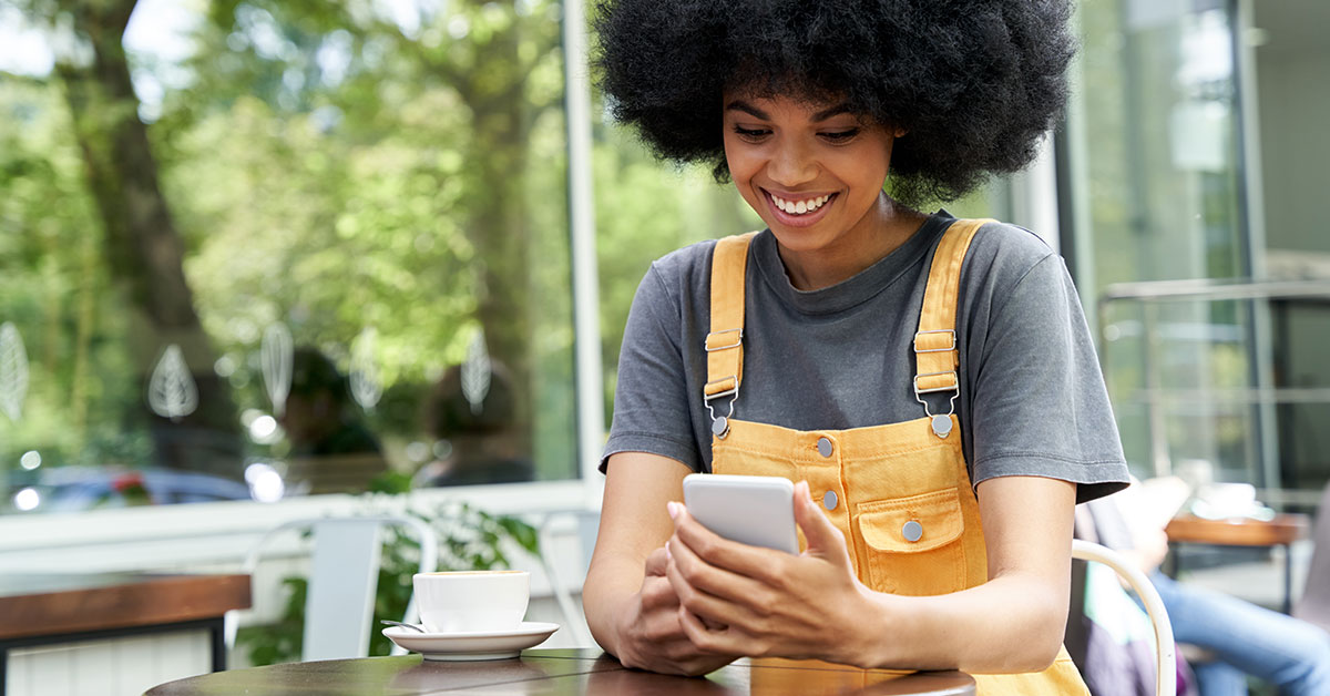 A teenage girl sits outside a café at a table. She is looking down at her phone in her hands and smiling.