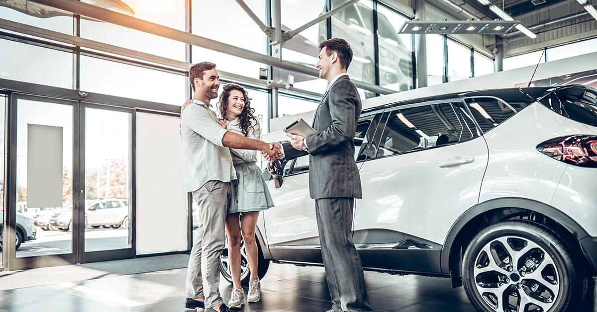 Young couple inside a car dealership next to a white SUV. They are hugging, smiling, and shaking hands with a car salesman in a suit.