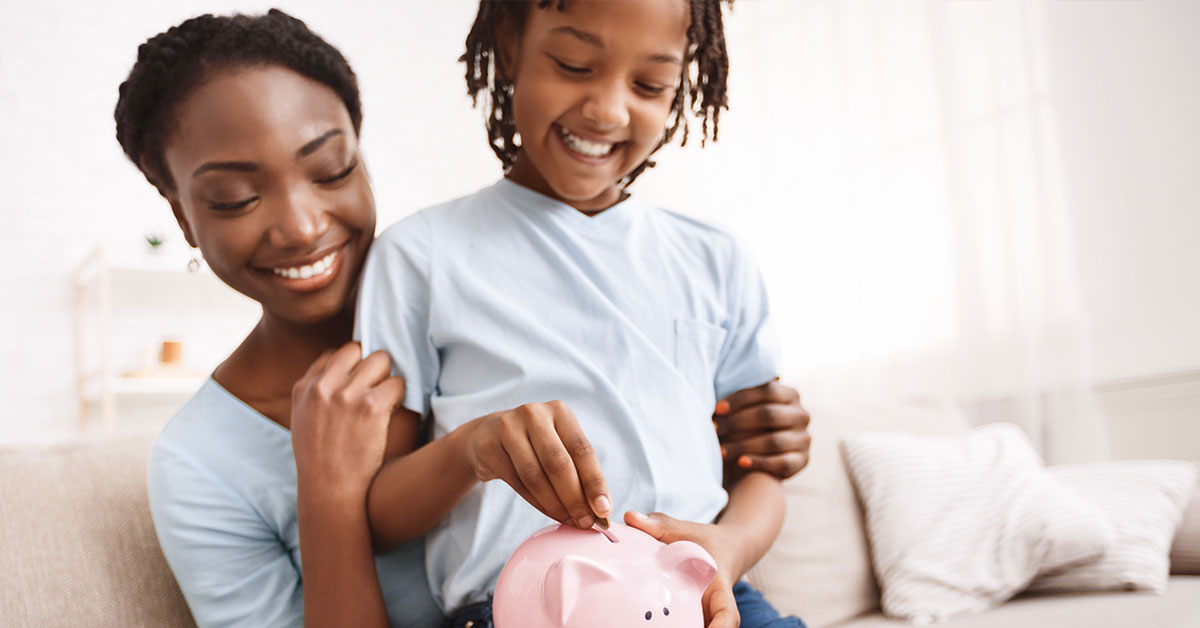 A young child is sitting on their mother’s lap, smiling and putting money into a piggy bank.