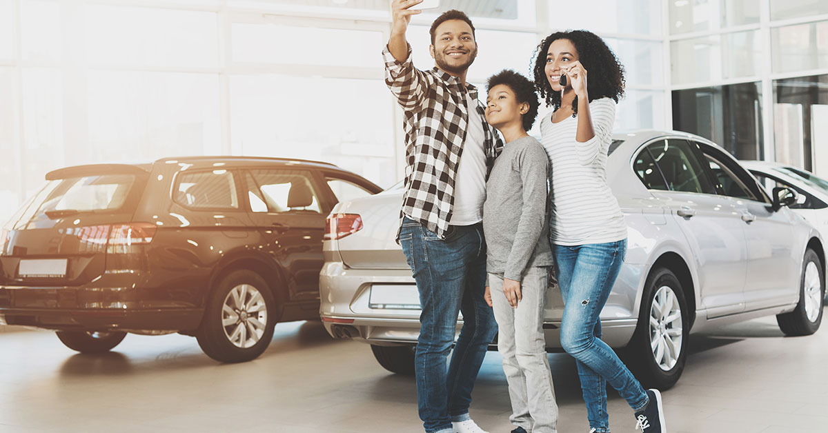 A young family stands in the showroom of a car dealership. The father is taking a selfie of the family in front of one of the cars.