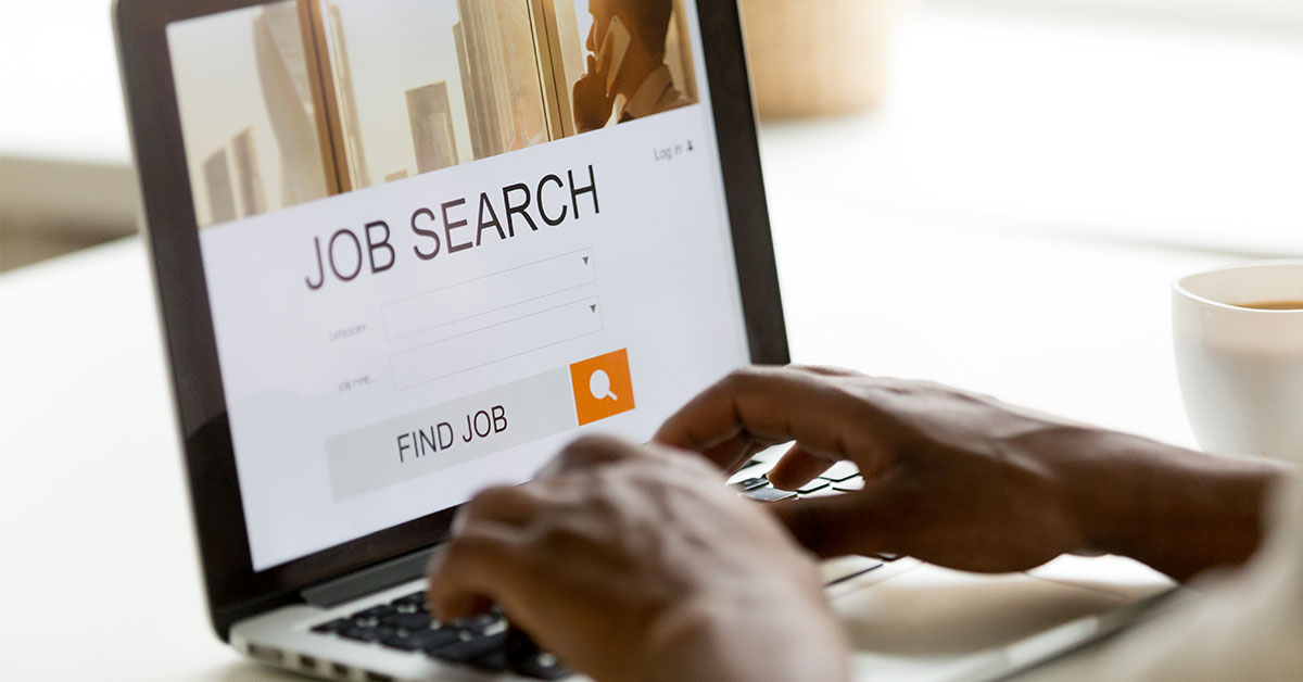 Close up of a man’s hands typing on a laptop. On the screen, we see a job search website.