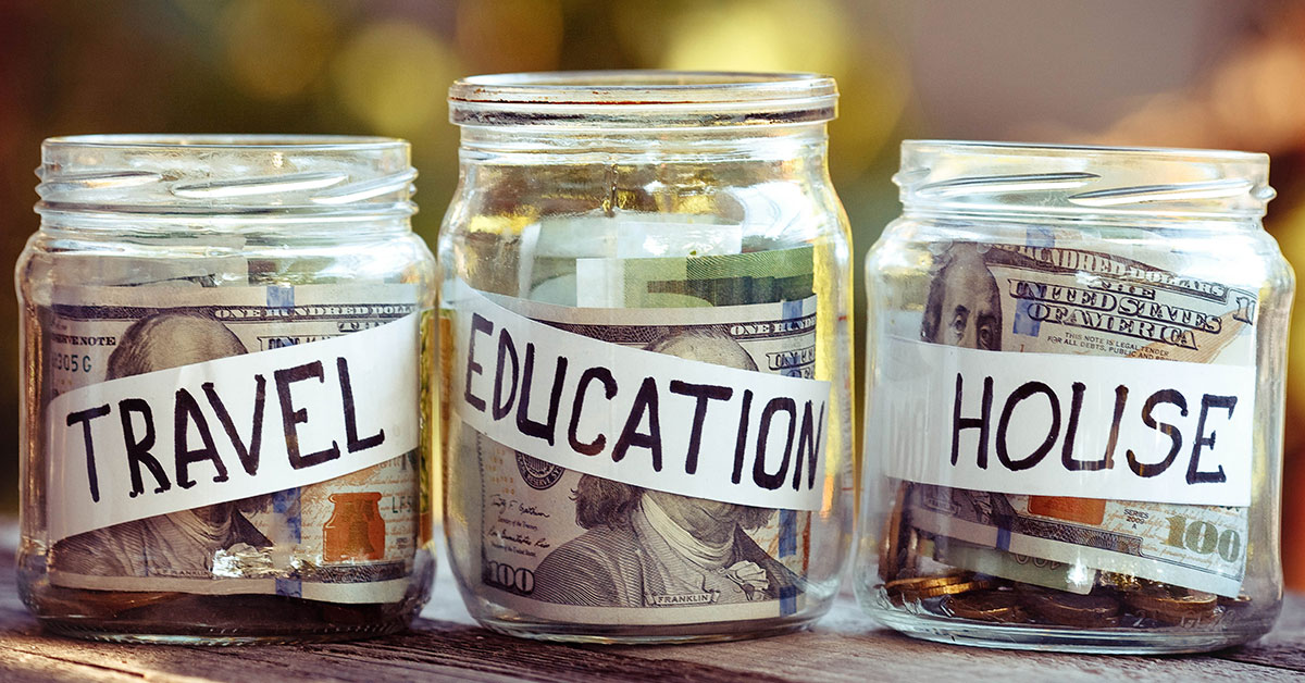 Three savings jars labeled 'Travel,' 'Education,' and 'House' sit on top of a table outside with loose money in them.