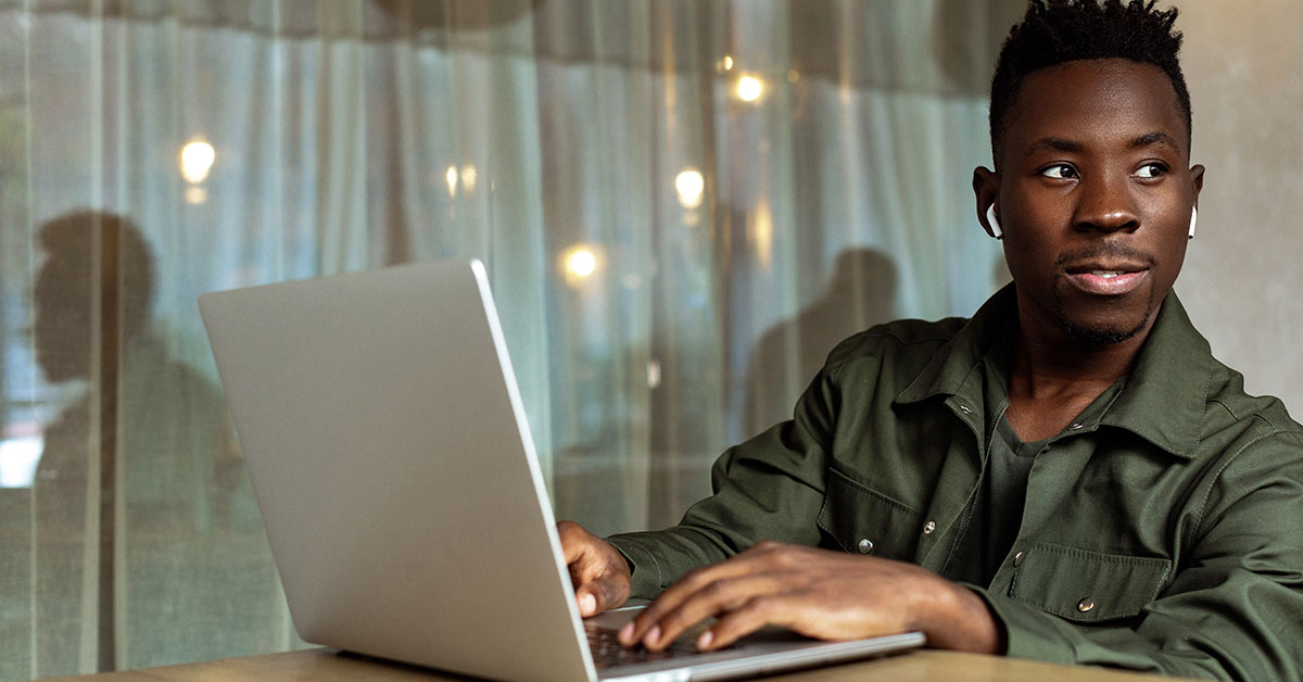 A young man sits outside at a table with his laptop. He has wireless earbuds in his ears and is looking off into the distance.